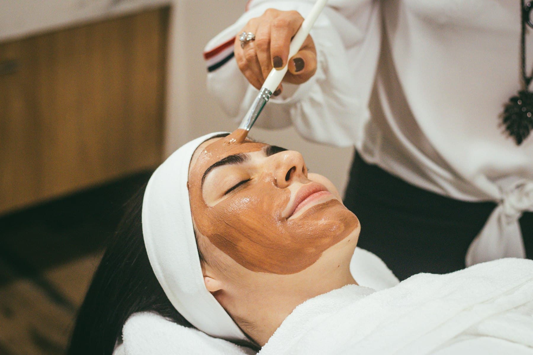 Kvinna som vilar under en ansiktsmask medan behandlaren penslar på produkten